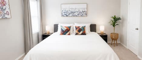 A bedroom with a bed, two nightstands, a ceiling fan, a window with curtains, a wall-mounted artwork, and a potted plant.
