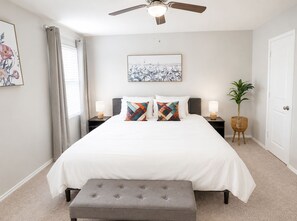 Een slaapkamer met een bed, twee nachtkastjes, een plafondventilator, een raam met gordijnen, een kunstwerk aan de muur en een bloempot met plant.