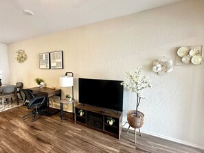 Una sala de estar con un televisor de pantalla plana, una mesa de comedor, sillas, una mesa de centro, una lámpara y adornos en la pared.