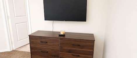 A room with a dark wooden dresser, a flat-screen TV mounted on the wall, and a small table with a candle.