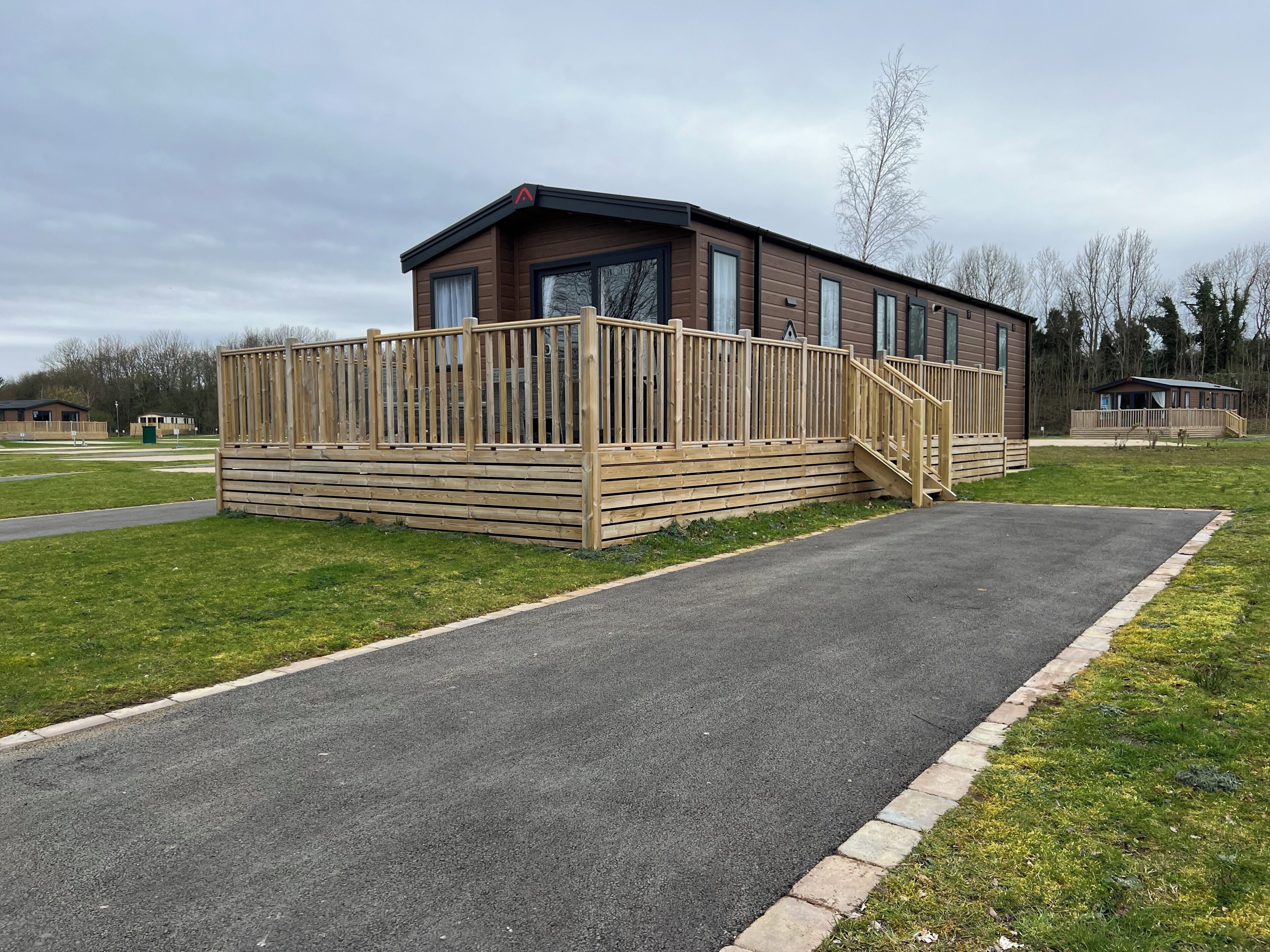 A single-story wooden building with a fenced deck, a paved driveway, and a grassy area.