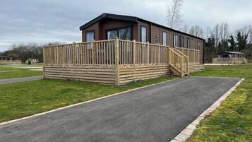 A single-story wooden building with a fenced deck, a paved driveway, and a grassy area.