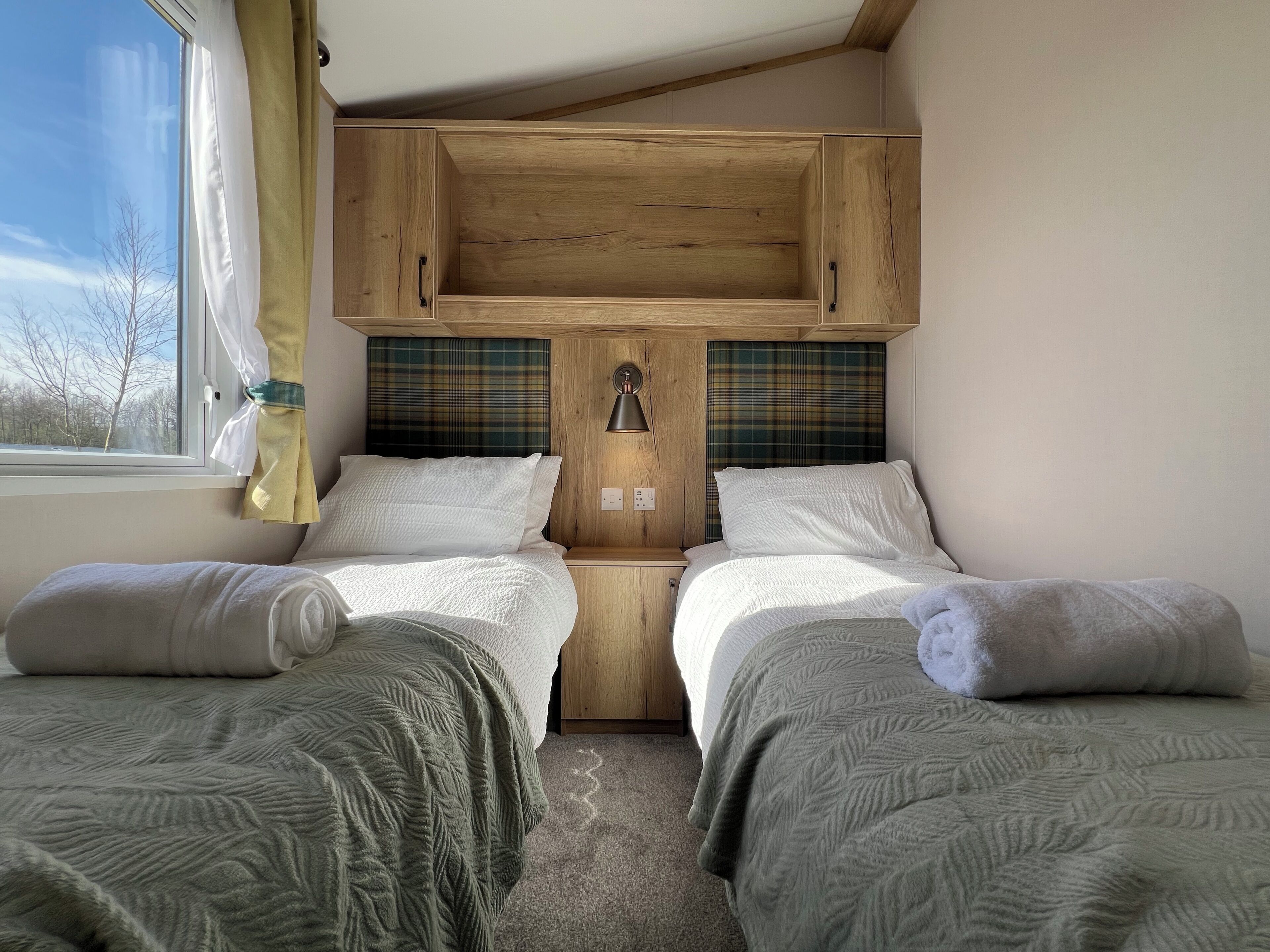 Ein Zimmer mit zwei Betten, Holz-Kopfteilen, einem Fenster mit Vorhängen und einer Wandlampe.