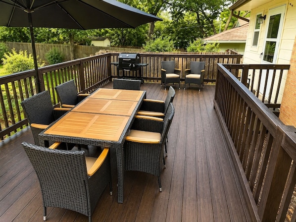 Una terraza con mesa de madera, sillas de mimbre, una parrilla y una zona cubierta con sombrilla.