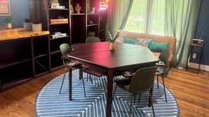A dining area with a wooden table, chairs, a blue patterned rug, and a bookshelf with various items.