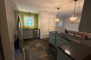 A kitchen with a refrigerator, a sink, a window with blinds, a door, and pendant lights.