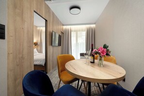 Ein Hotelzimmer mit einer Wandpaneele aus Holz, einem runden Holztisch mit Stühlen, einem Bett mit weißer Bettwäsche, einem angebrachten Fernseher und einem Fenster mit Vorhängen.