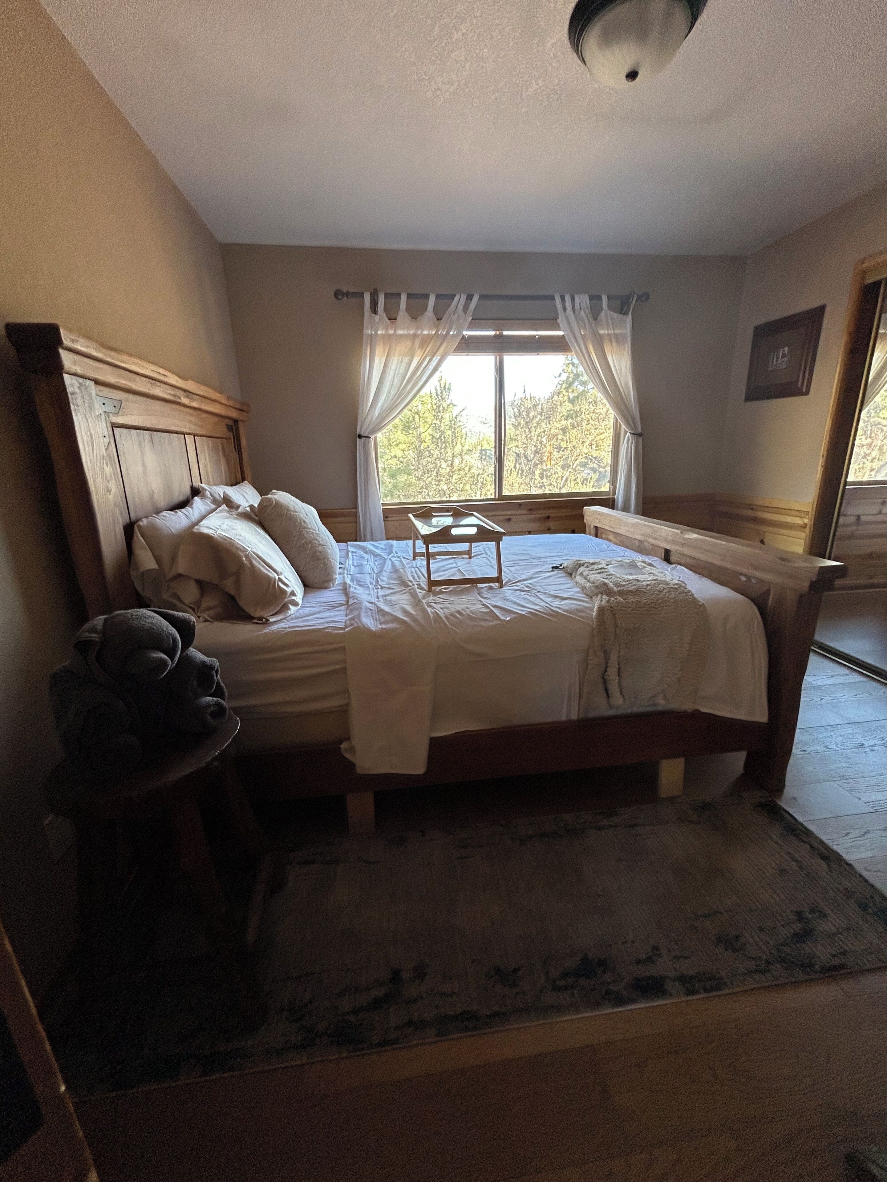 A bedroom with a wooden bed, a bedside table, a nightstand, a window with curtains, and a view of trees outside.