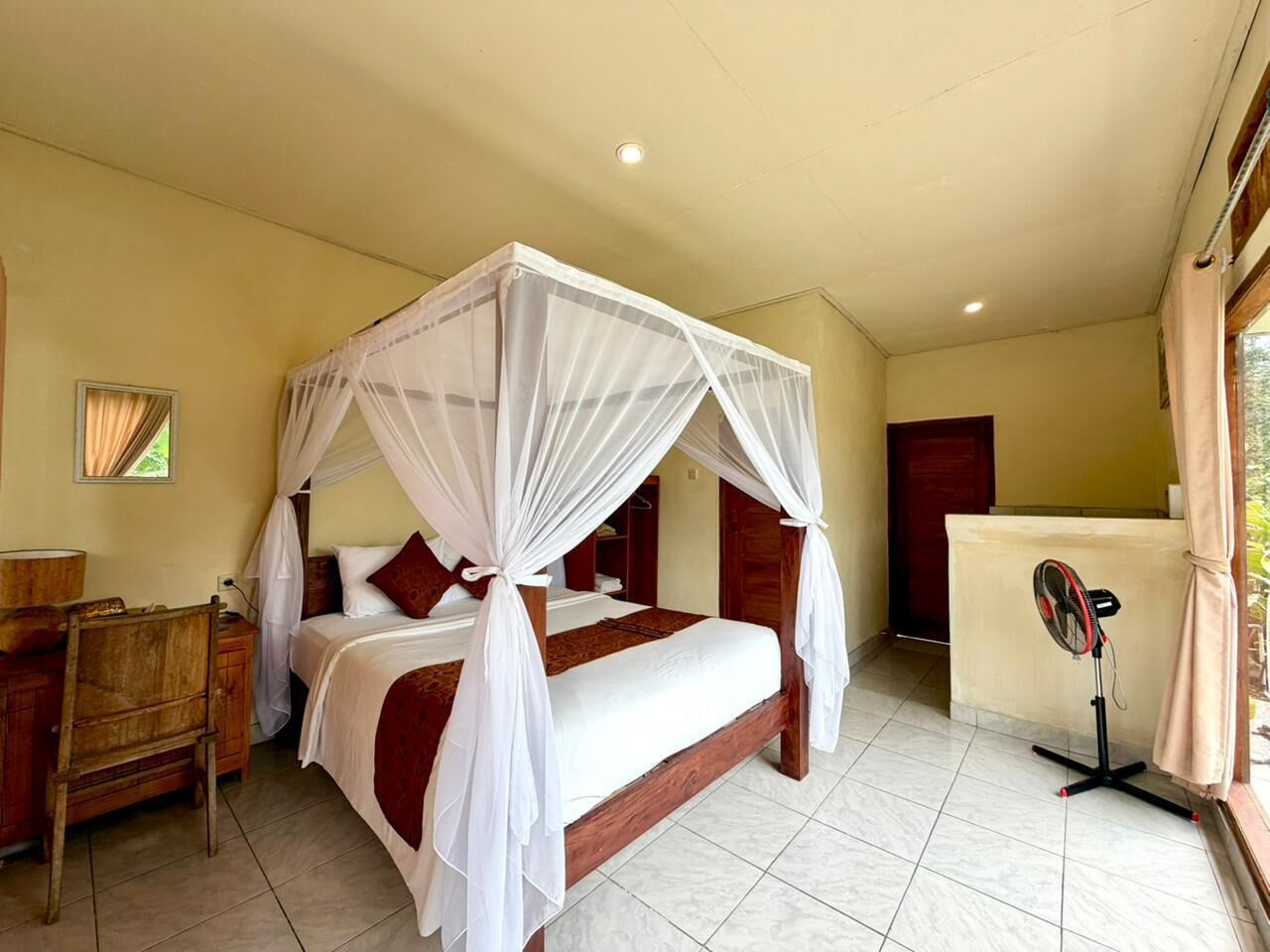 A single bed with a canopy, a wooden nightstand, a fan, and a window with curtains.
