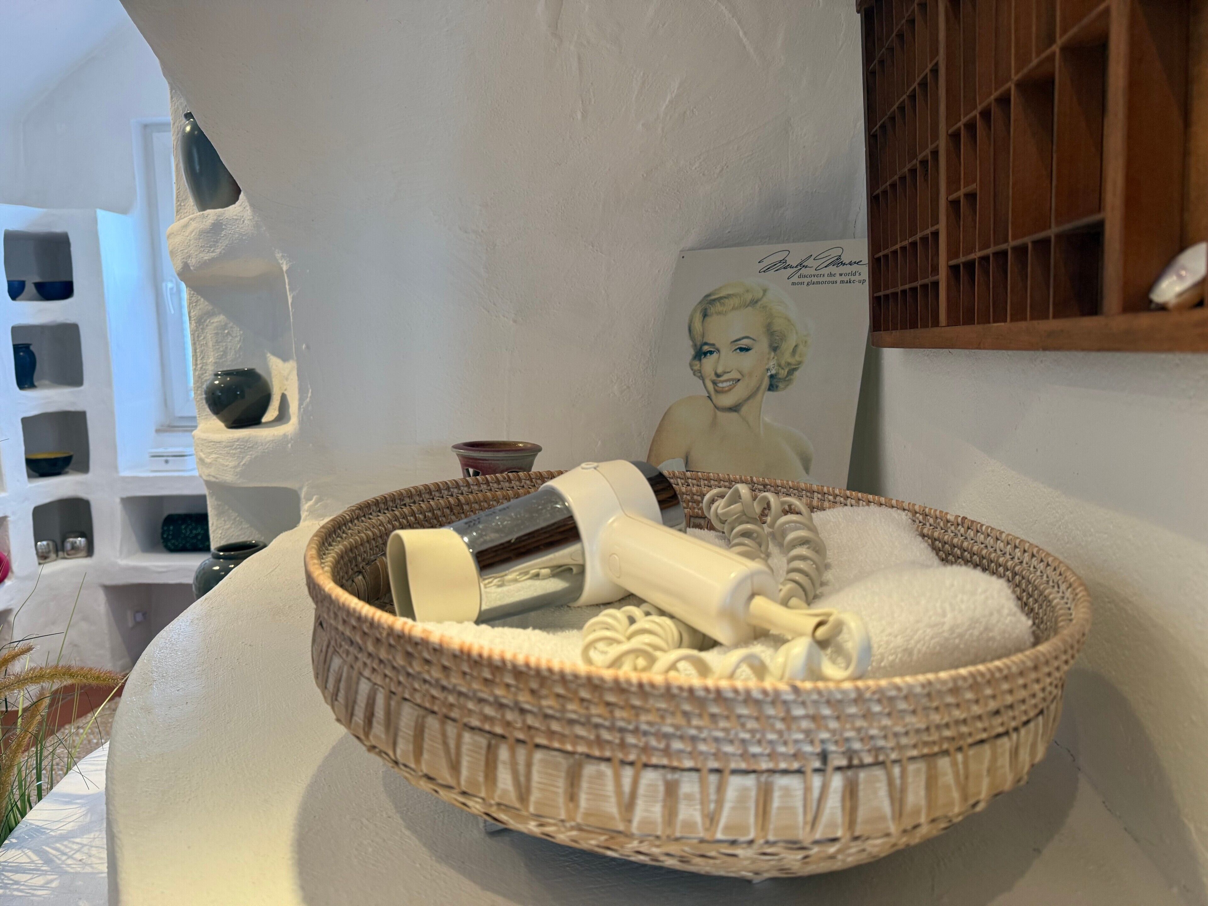 A room with a white wall, a poster of Marilyn Monroe, a wicker basket with toiletries, and a white shelf with decorative items.