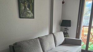 A grey sofa in a room with a painting on the wall, a lamp, and a view through a glass door. - Cocon Neuf & Vue Panoramique - Portes du Soleil (Vionnaz)