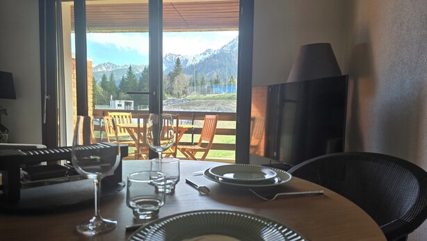 A dining table with plates, glasses, and cutlery, a TV, and a view of mountains through a glass door. - Cocon Neuf & Vue Panoramique - Portes du Soleil (Vionnaz)