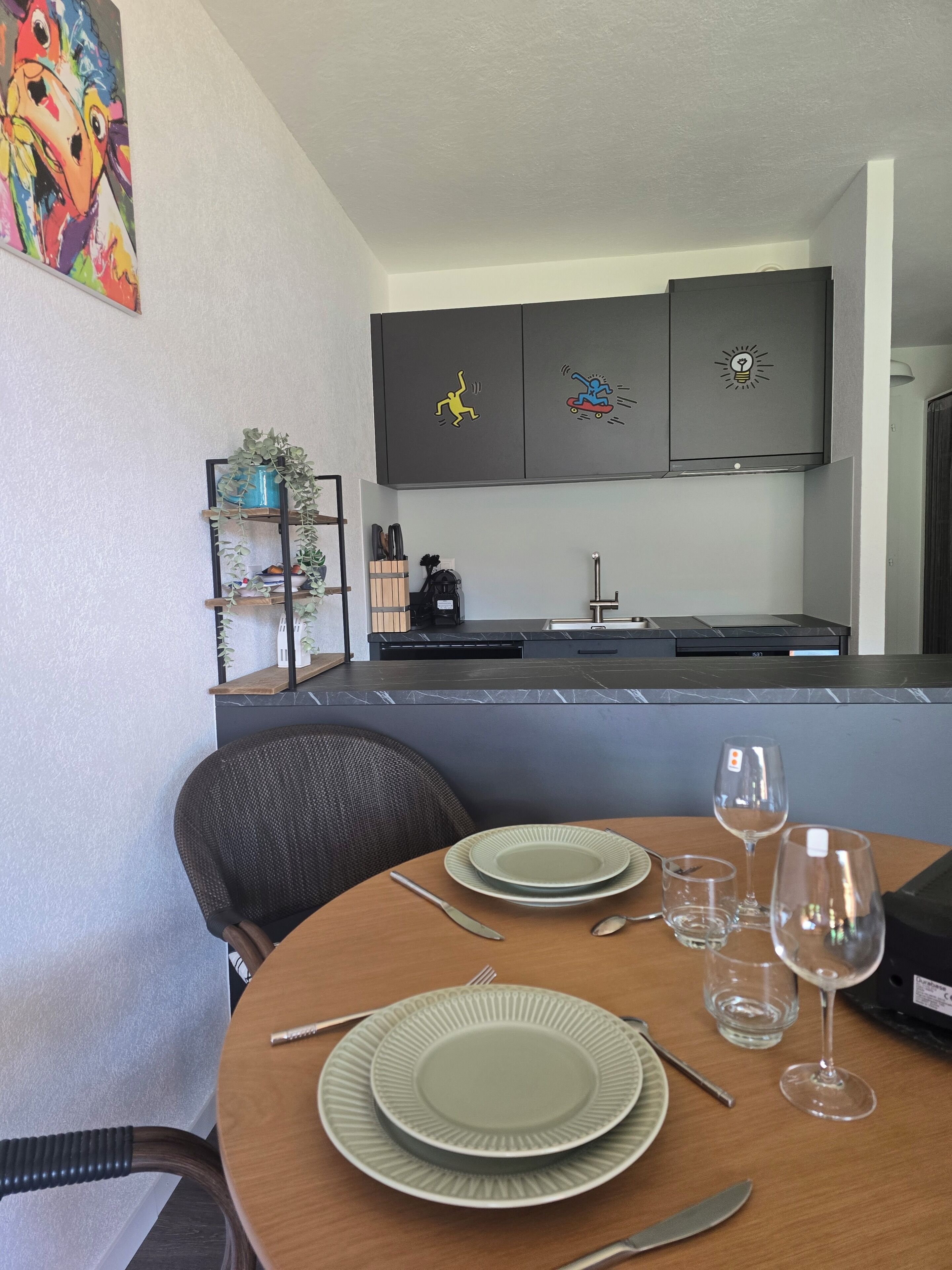 A dining area with a table set for two, a chair, a painting on the wall, and a kitchen in the background.
