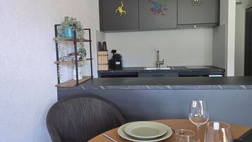A dining area with a table set for two, a chair, a painting on the wall, and a kitchen in the background.