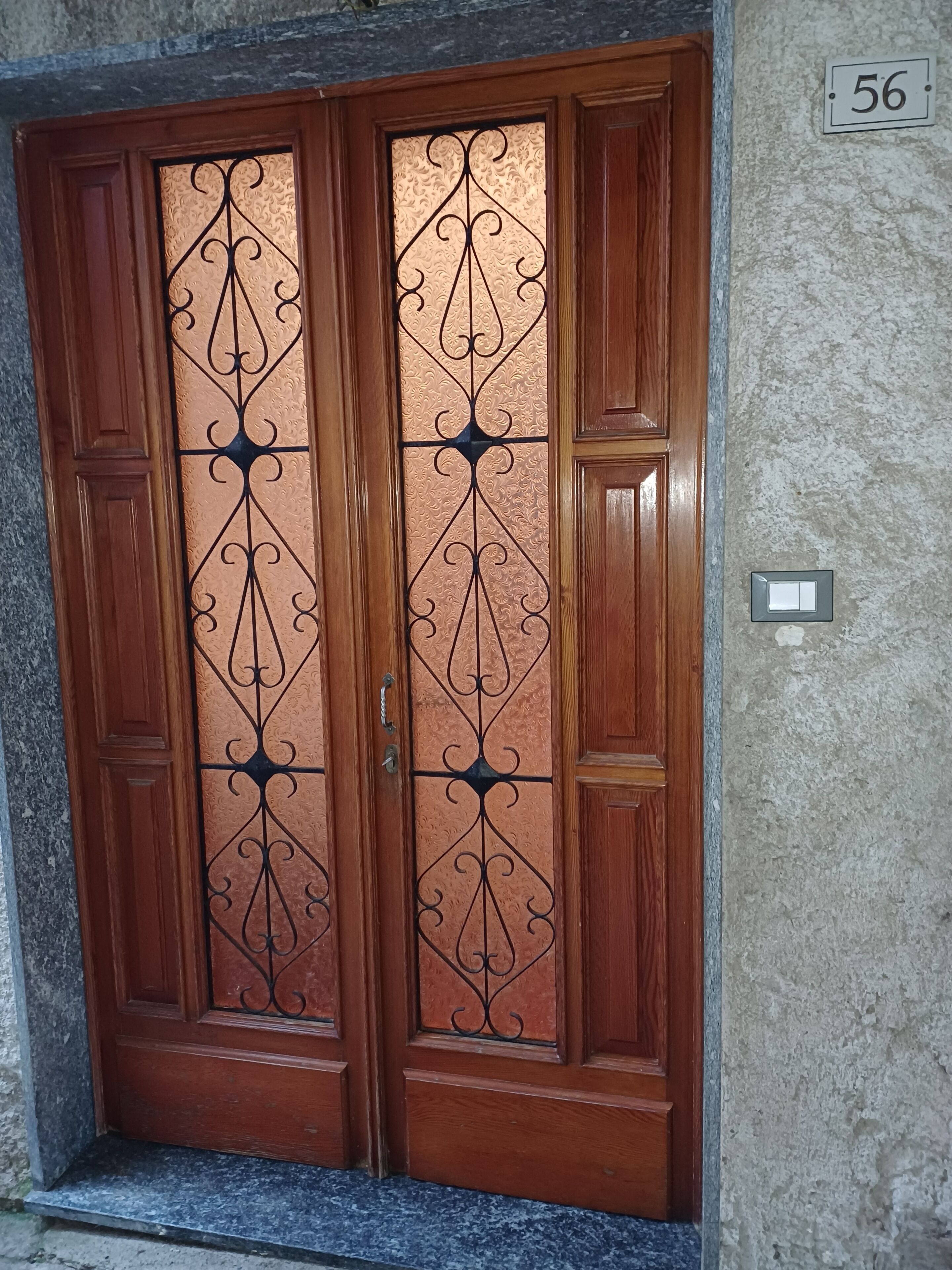 Una porta doble de fusta amb treball metàl·lic detallat, una paret de pedra i el número 56.