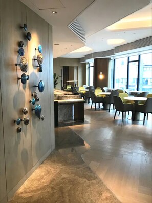 Un intérieur moderne agrémenté d’un élément décoratif fixé au mur, d’un espace salon avec tables et chaises, et d’une grande fenêtre laissant entrer la lumière naturelle.