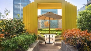 Une structure jaune avec une table et un parasol, entourée de verdure et de b âtiments.
