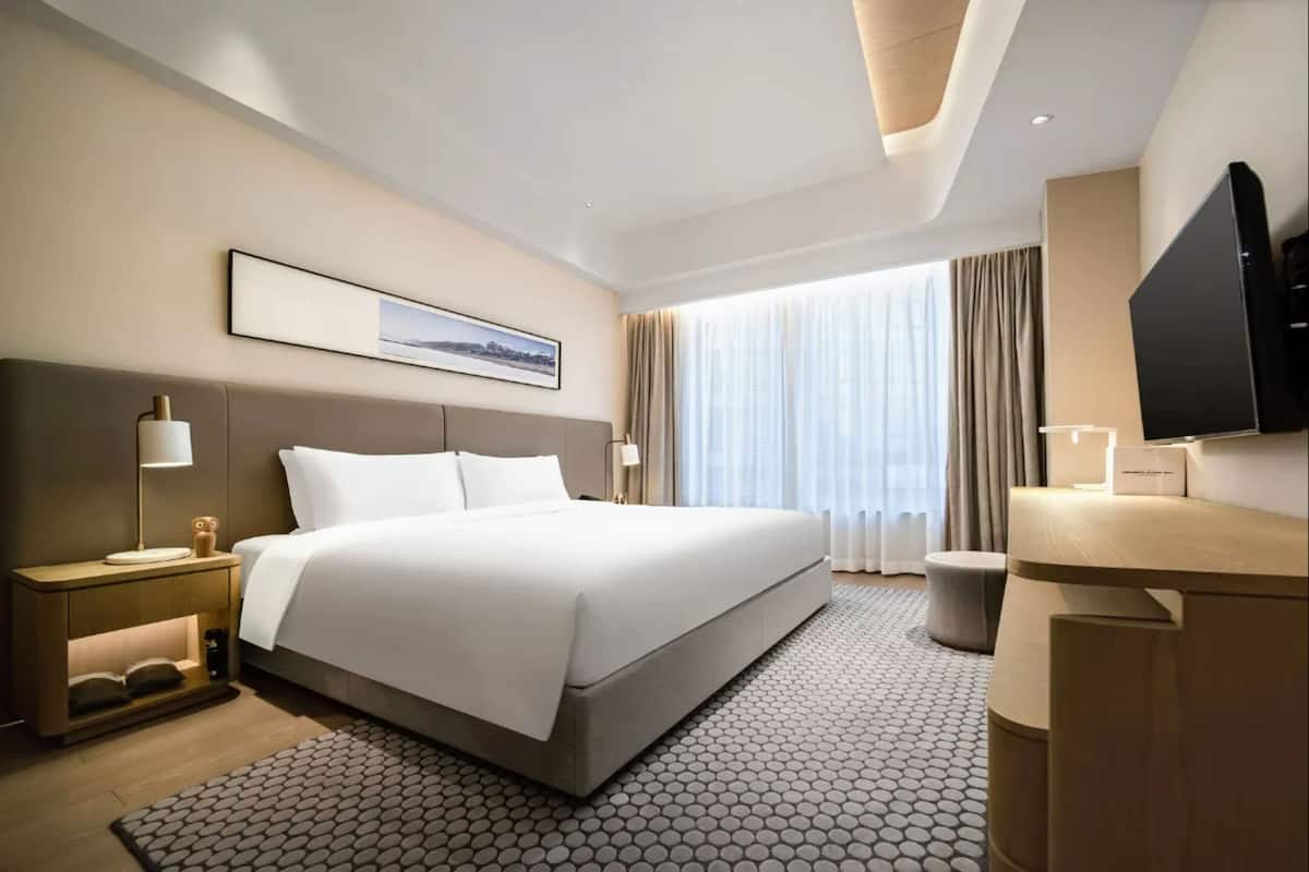 Une chambre d’hôtel avec un grand lit, un bureau doté d’un téléviseur à écran plat, une table de chevet avec une lampe et une fenêtre avec des rideaux.