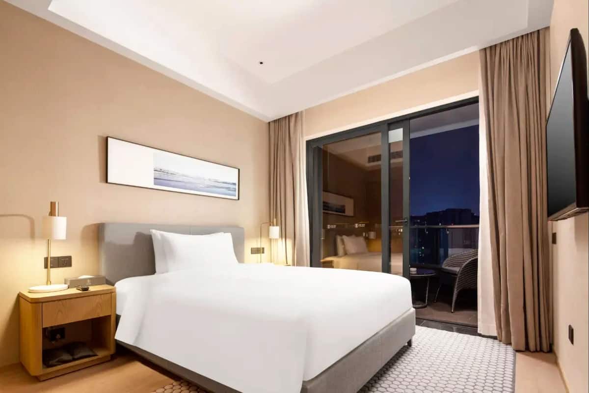 Une chambre d’hôtel comprenant un lit, des tables de chevet, un téléviseur à écran plat, une porte-fenêtre coulissante donnant sur un balcon et un tableau encadré au mur.