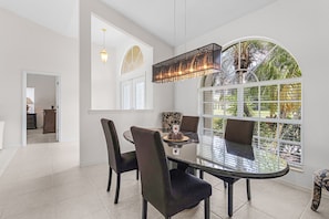 A dining area with a glass table, dark chairs, a hanging light fixture, and a view through a window with a white frame. - Villa Twin Palms 3BR 2BA Waterfront Heated Pool Gulf Access Private Dock (Cape Coral)