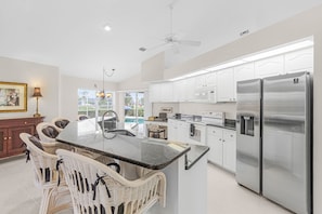 A spacious kitchen with a central island, white cabinetry, stainless steel appliances, and a dining area with wicker chairs. - Villa Twin Palms 3BR 2BA Waterfront Heated Pool Gulf Access Private Dock (Cape Coral)