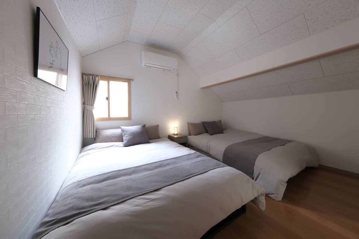 A bedroom with two beds, a window, a wall-mounted air conditioner, and a bedside table with a lamp.