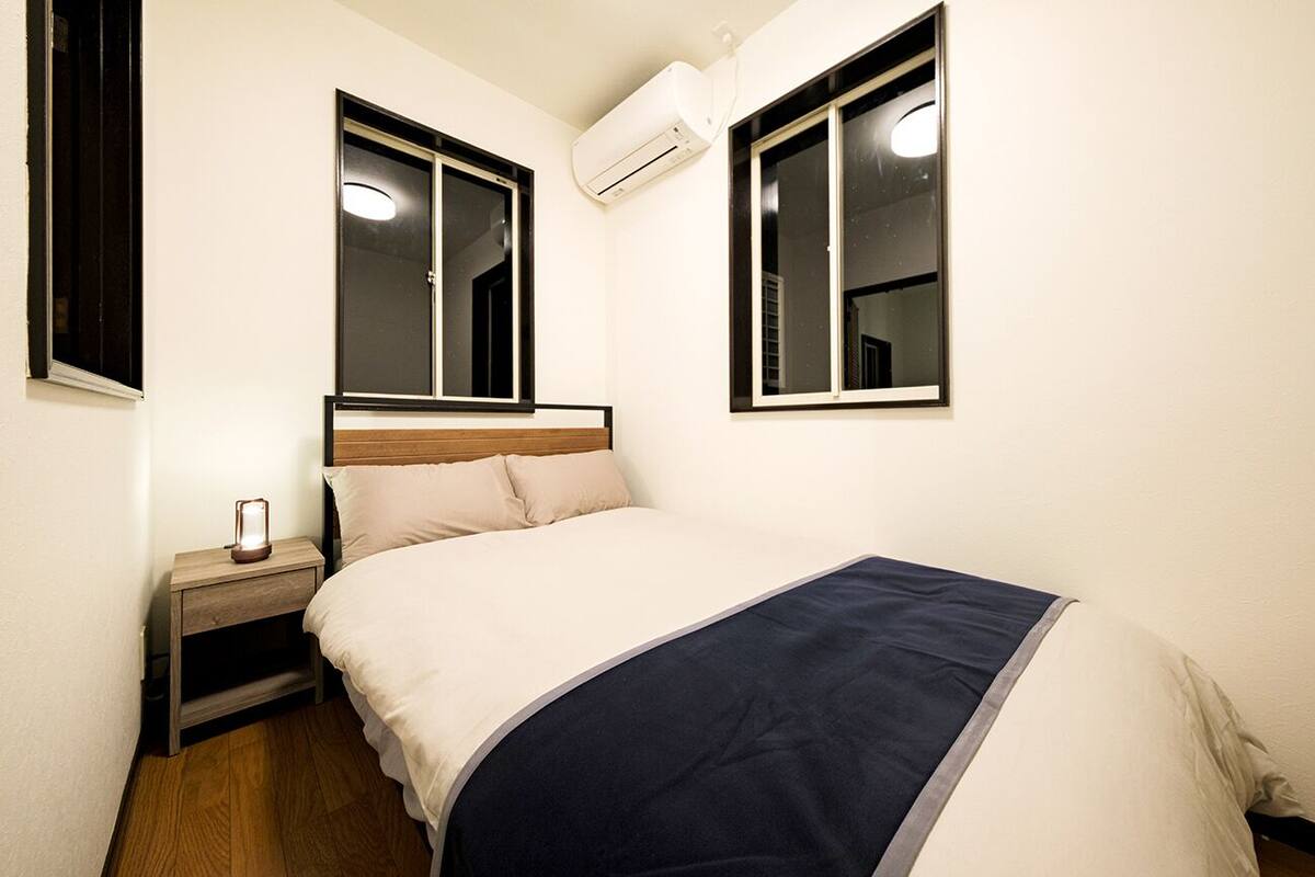 A bedroom with a bed, bedside table, nightstand, and two windows with air conditioning units.