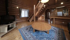 Ein Holzblockhaus-Innenraum mit Holzboden, einem runden Tisch, einem Fernseher auf einem Schrank, einer Treppe und einer Küchenzeile.
