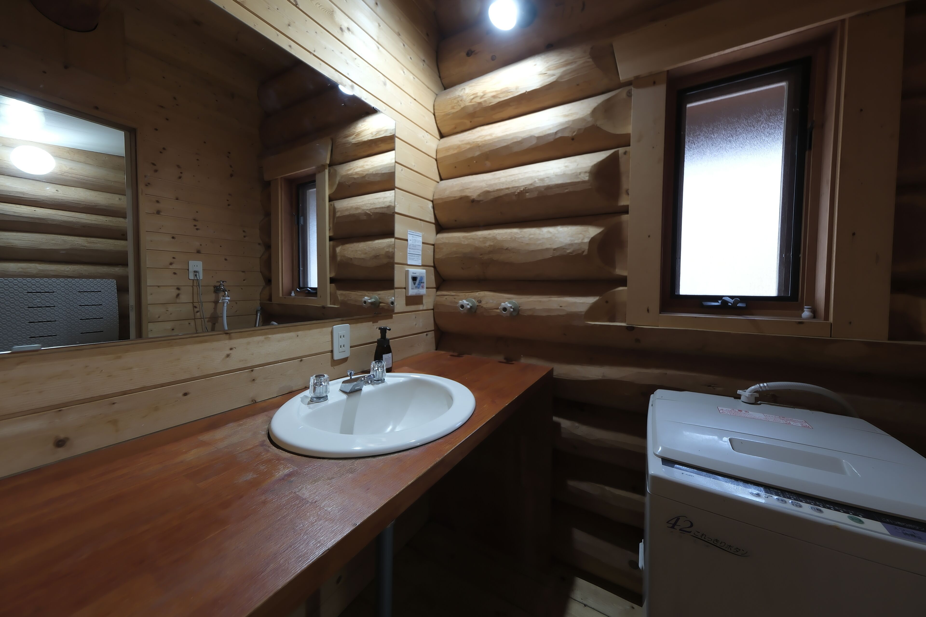 A wooden cabin-style bathroom with a wooden vanity, a sink, a mirror, a window, and a white portable heater.