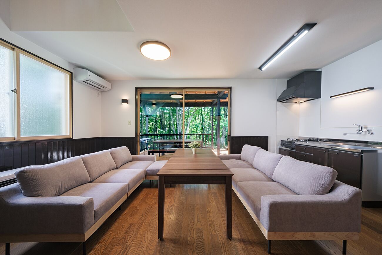 Una sala moderna con un sofá, una mesa de centro de madera, una zona de cocina y un ventanal con vistas a la vegetación.