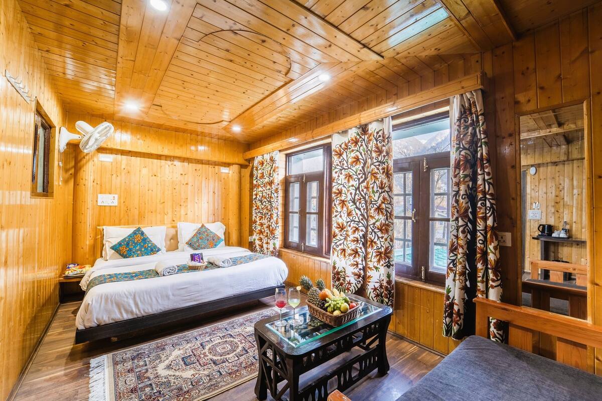 A wooden cabin room with a bed, a fan, a table with fruit, and patterned curtains.