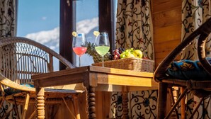 A wicker chair, a wooden table with a wicker basket, and a window with a view of the sky and clouds. - Bombay Palace (Pahalgam)