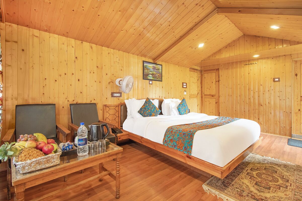 Una habitación con paneles de madera, una cama, un escritorio con una cesta de frutas, un ventilador y una cafetera.