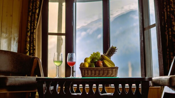 A room with a wooden bench, a table with a fruit basket and two glasses of wine, and a window with a mountain view. - Bombay Palace (Pahalgam)