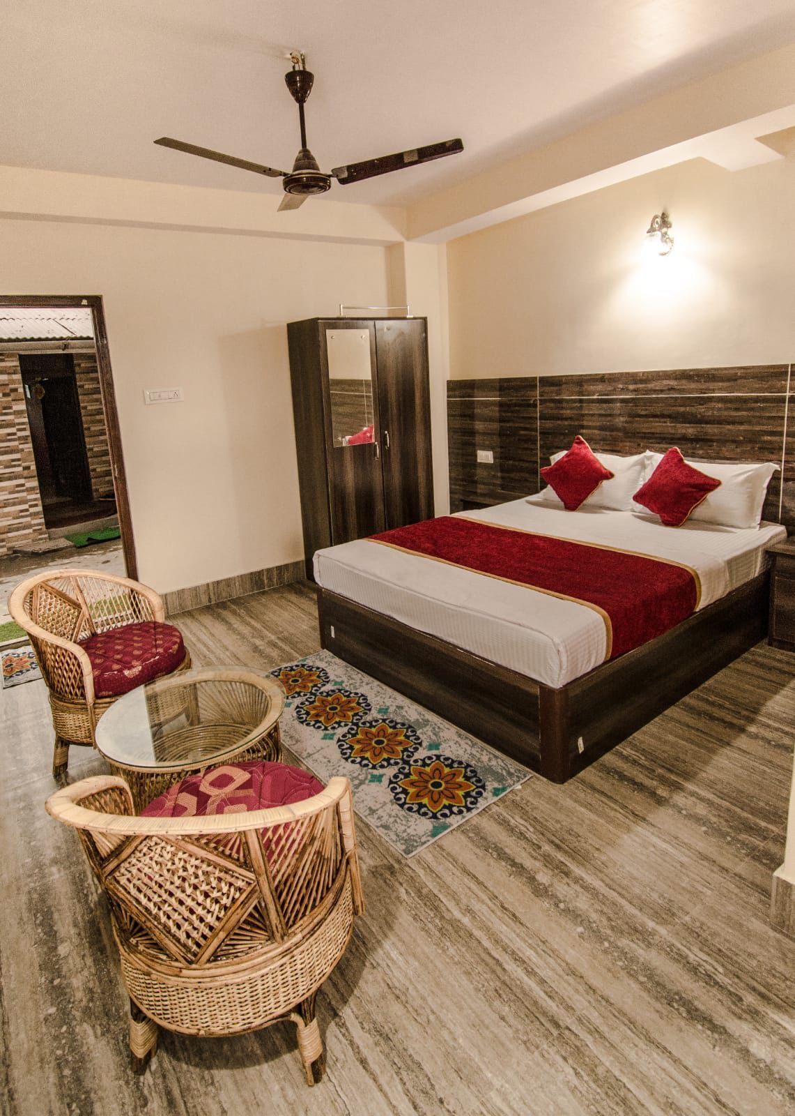 A hotel room with a bed, two wicker chairs, a small table, a ceiling fan, a wardrobe, and a patterned rug.
