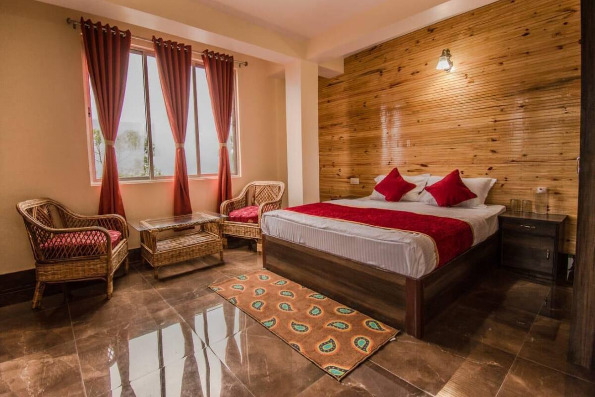 A bedroom with a wooden headboard, a bed with red pillows, a wicker chair, a small table, and a window with curtains.