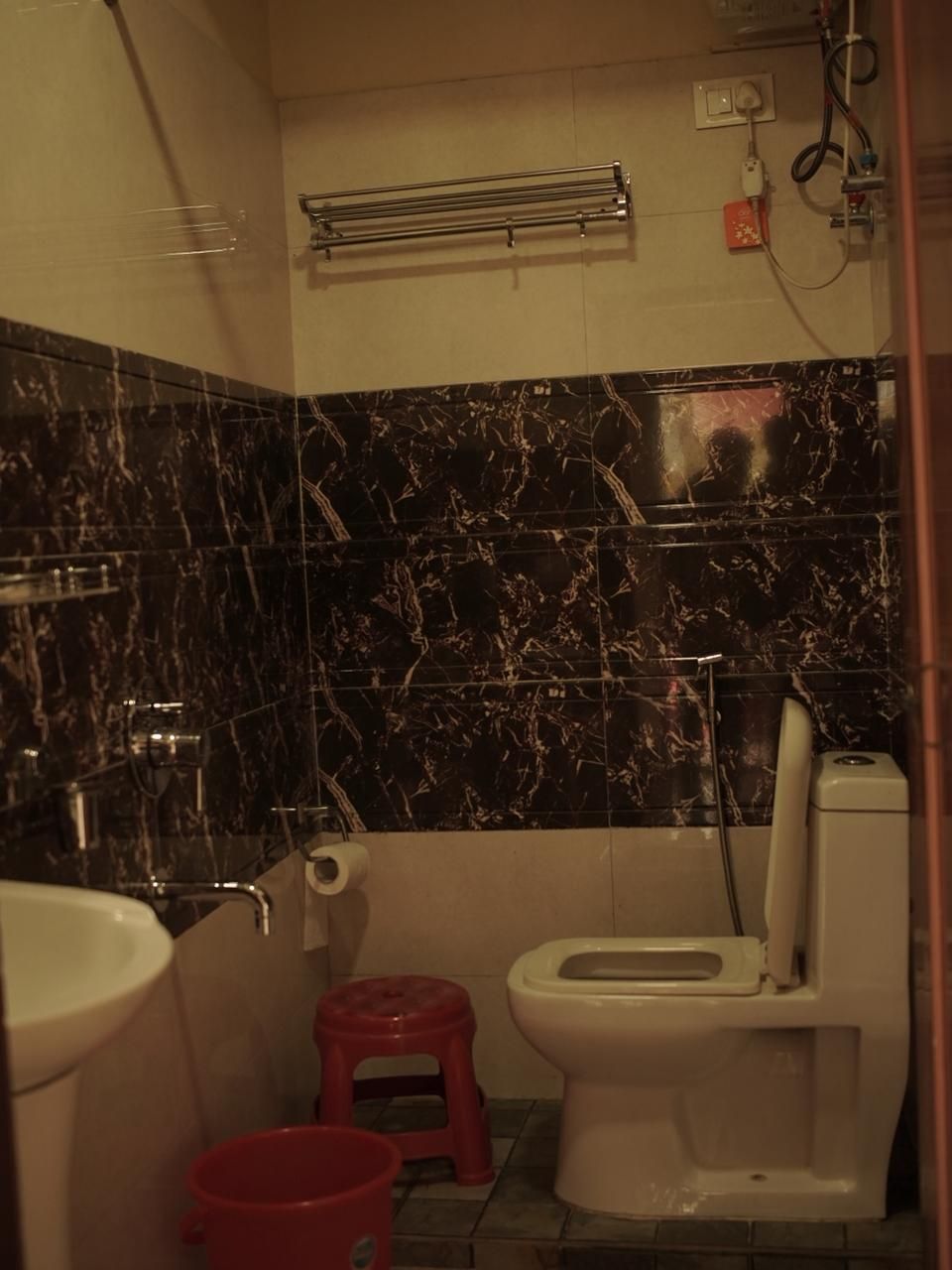 Ein Badezimmer mit Toilette, einem roten Hocker, einem roten Eimer, einem Handtuchhalter und einer Duschbrause.