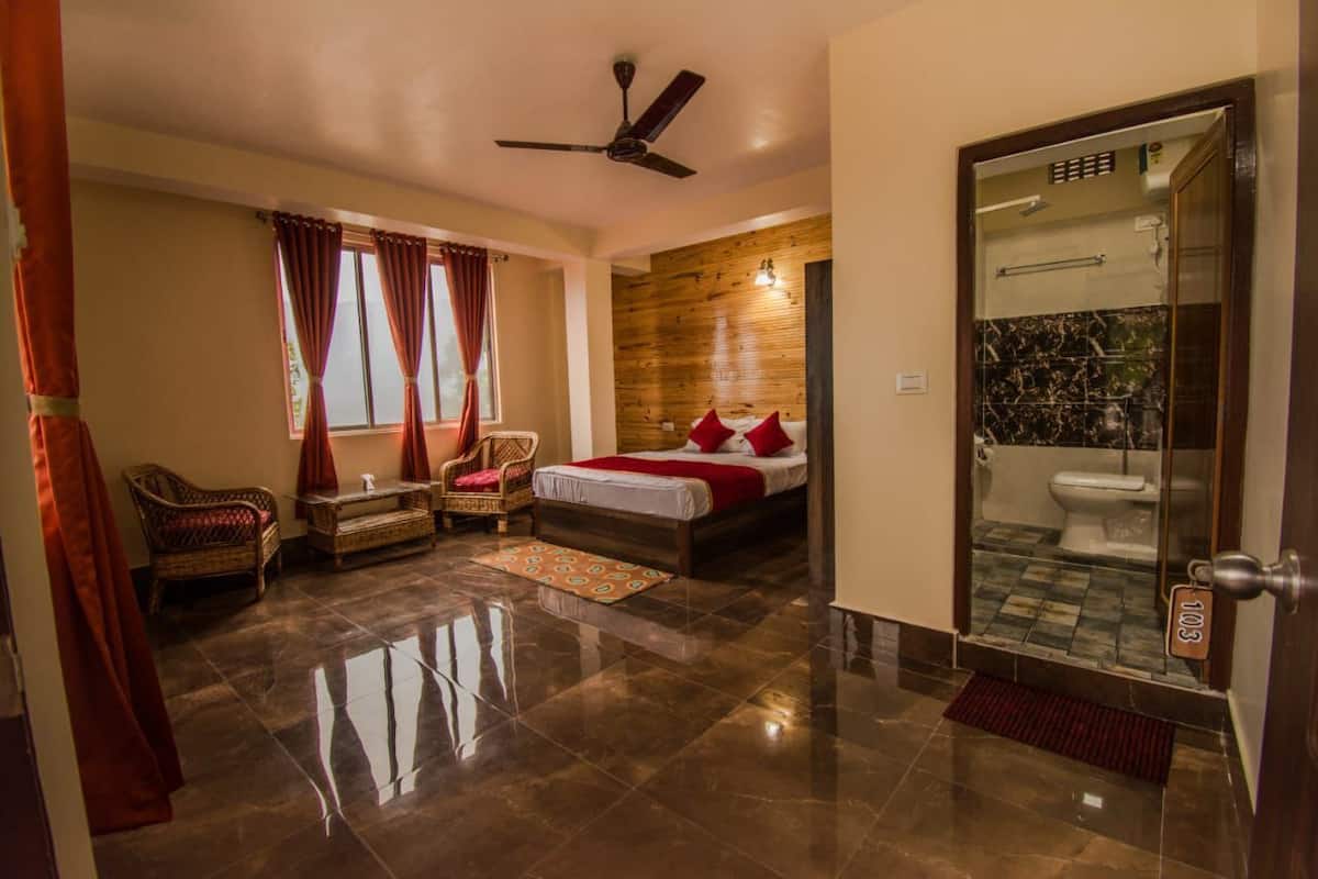 A hotel room with a bed, a sitting area with a chair and a small table, a ceiling fan, a bathroom with a glass door, and a tiled floor.