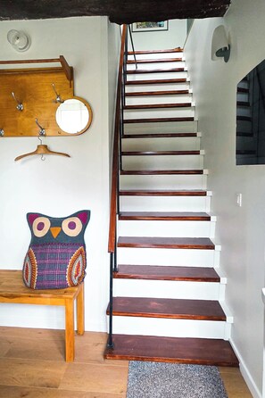 Una escalera de madera con peldaños y barandilla, un banco de madera con un cojín de búho de colores, un perchero de madera con ganchos y un espejo fijado a la pared.