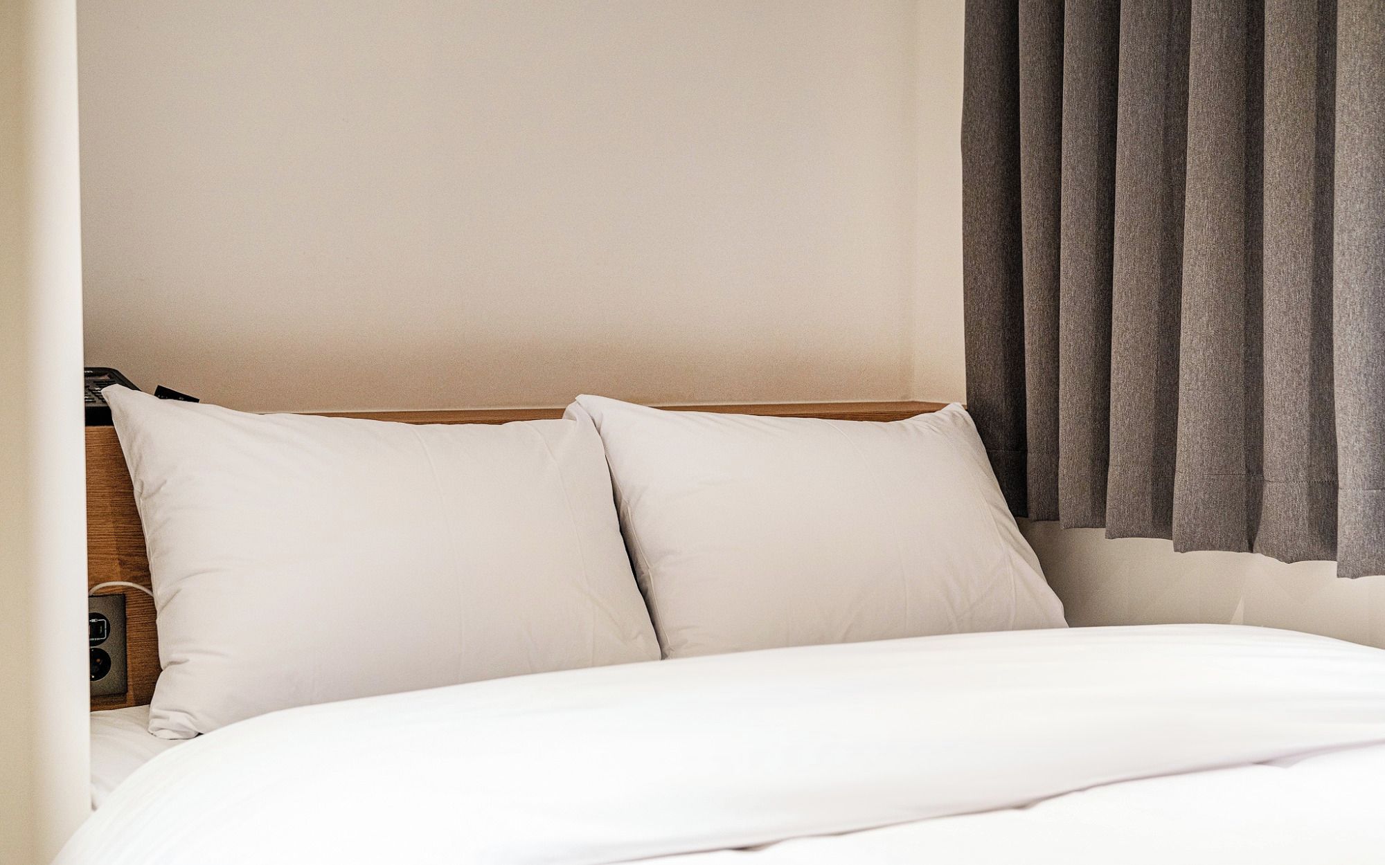 Una habitación de hotel con cama, dos almohadas, cabecero de madera, toma de corriente y ventana con cortinas.