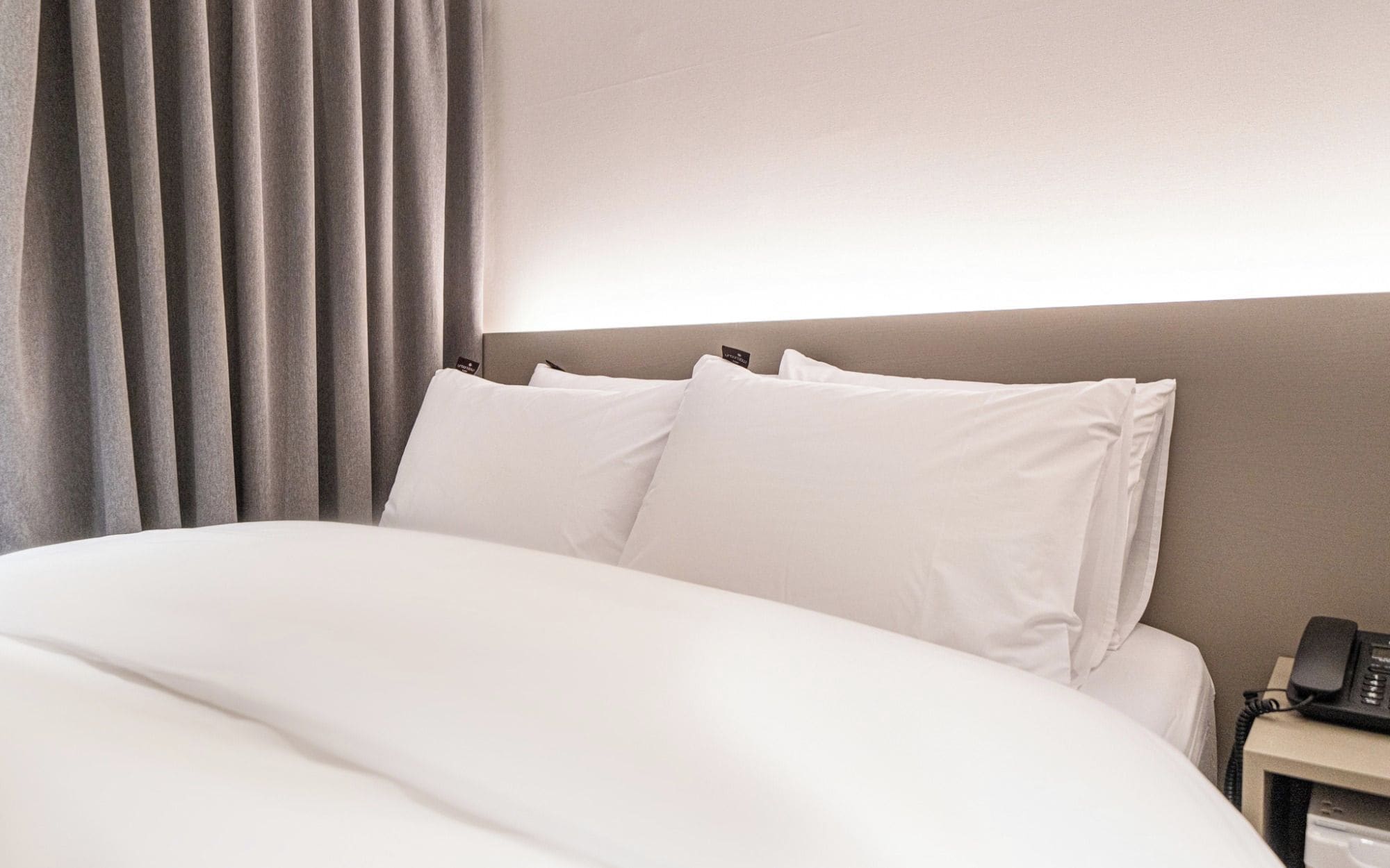 A hotel room with a bed, pillows, a nightstand with a phone, and a curtain.