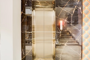 An elevator with a metallic finish, a glass door, and a reflection of stairs and a light fixture. - Deluxe Palette Bareilly Bellazio Suites (Bareilly)