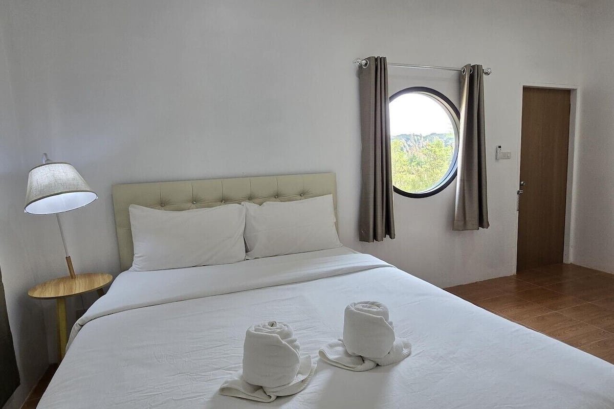 Une chambre d’hôtel avec un lit, une fenêtre ronde, des rideaux, une lampe et des serviettes sur le lit.