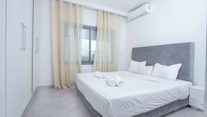 Ein modernes Schlafzimmer mit einem großen Bett, einem grauen Kopfteil, weißer Bettwäsche und einem Fenster mit transparenten Vorhängen.