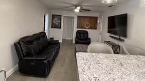 A living room with a black leather sofa, a dining table with chairs, a ceiling fan, and a TV mounted on the wall.