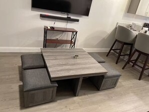 A modern living room with a grey sectional sofa, a wooden coffee table, a flat-screen TV mounted on the wall, and a kitchen area with bar stools.