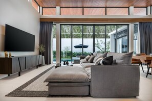 A modern living room with a large flat-screen TV, a sectional sofa, and a dining area with a table and chairs. - Space Botanica Modern Loft pool villa 29 Phuket-1 (Phuket)