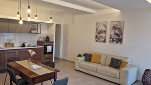 Una cocina moderna con encimeras de mármol, armarios de madera e iluminación colgante. Hay una zona de comedor con una mesa y sillas de madera, y un sofá beige con cojines.