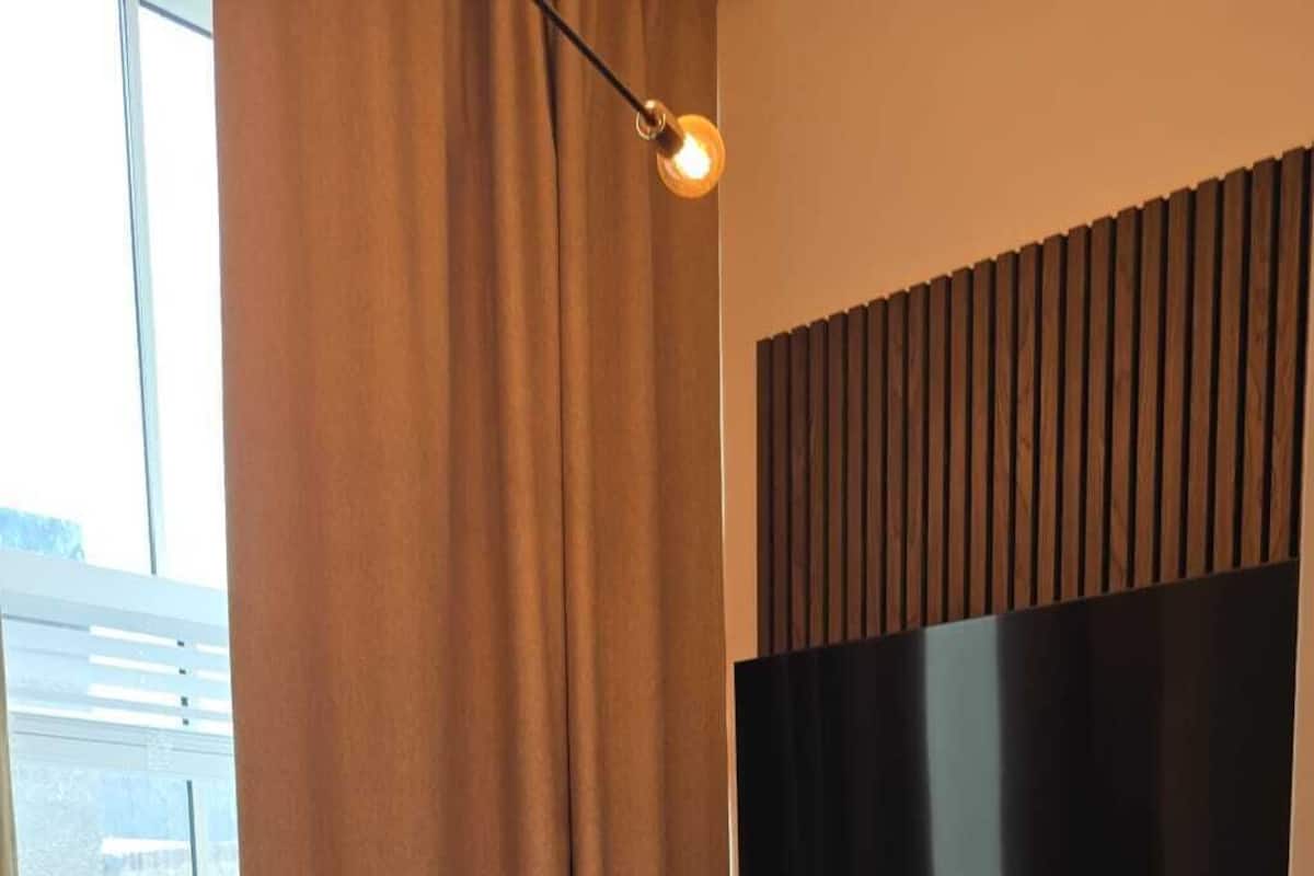 Habitación de hotel con una cama, un televisor de pantalla plana montado en una pared de paneles de madera y una ventana con cortinas.