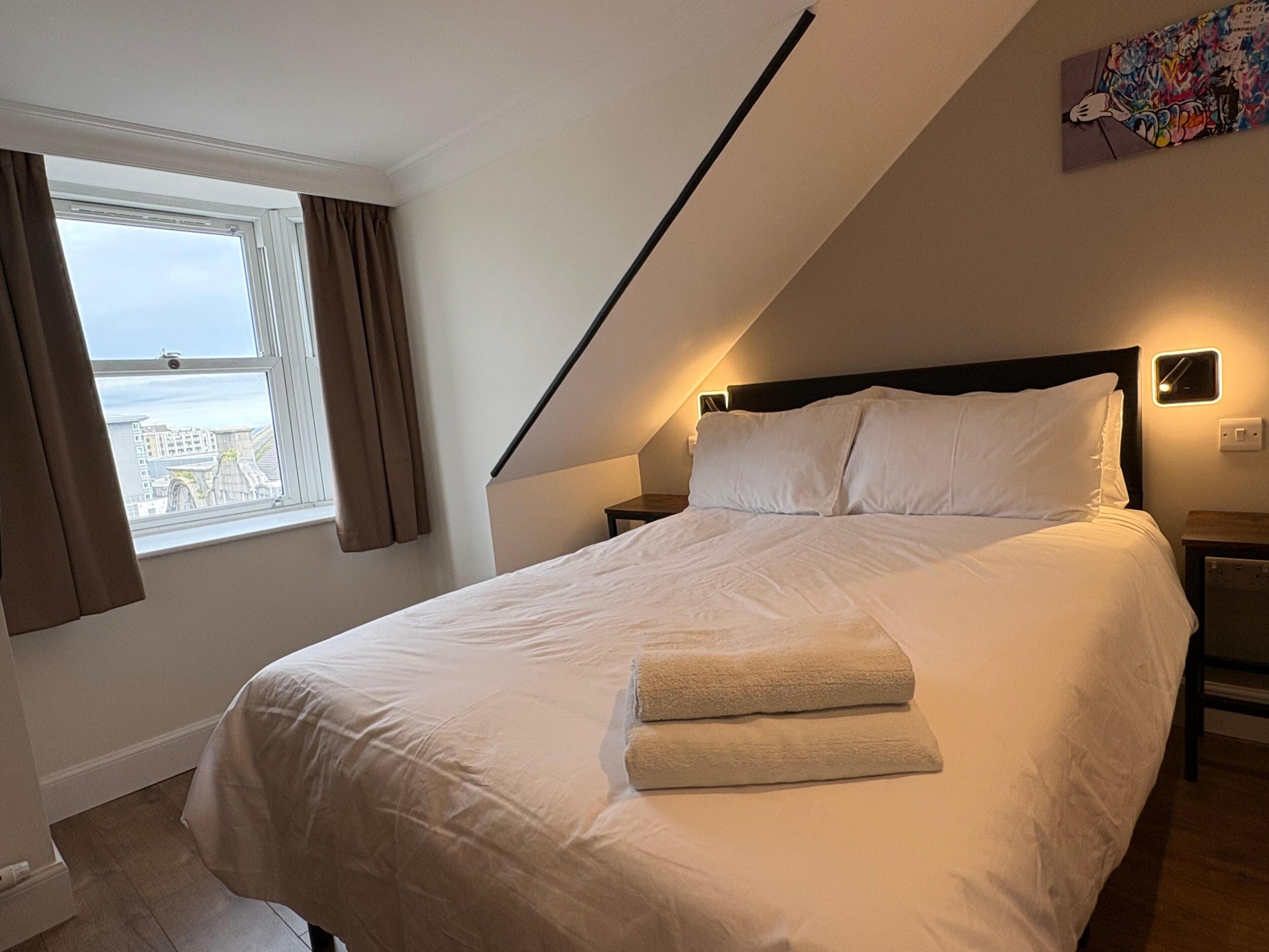 A bedroom with a bed, a nightstand, a window with curtains, and a view of the outside.